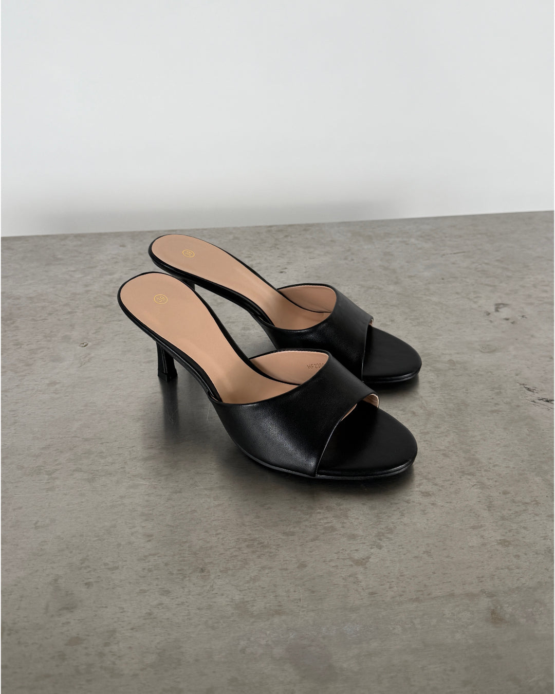 Sort højhælet sandal står stille på en betonoverflade. Sandalen har en enkel rem over foden, åben tå, og stilet hæl. Omgivelsen er minimalistisk med en lys baggrund.