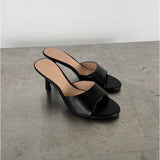 Sort højhælet sandal står stille på en betonoverflade. Sandalen har en enkel rem over foden, åben tå, og stilet hæl. Omgivelsen er minimalistisk med en lys baggrund.