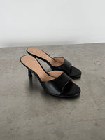 Sort højhælet sandal står stille på en betonoverflade. Sandalen har en enkel rem over foden, åben tå, og stilet hæl. Omgivelsen er minimalistisk med en lys baggrund.