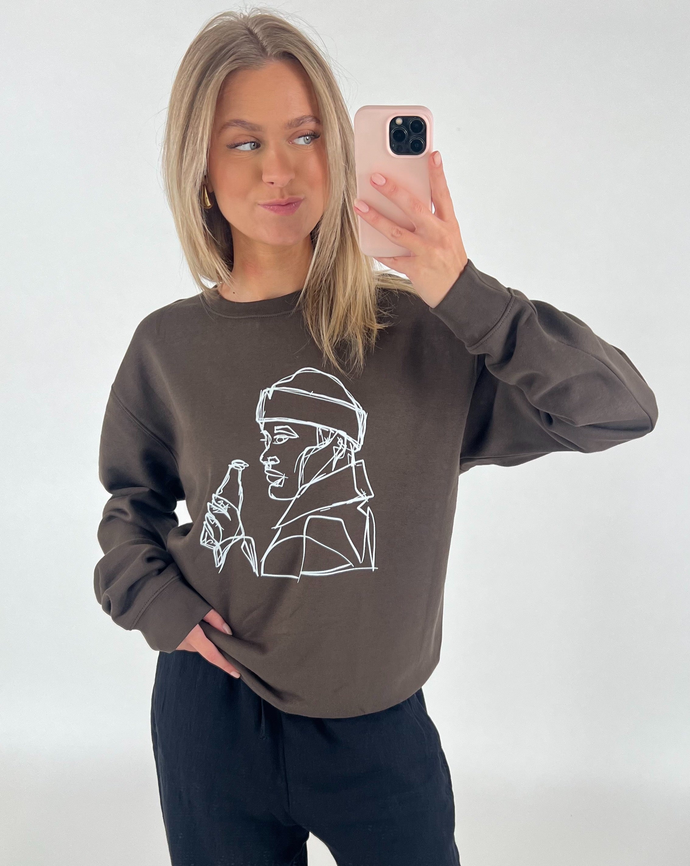 En person iført en brun trøje med hvid stregtegning tager en selfie med en mobiltelefon. Personen står foran en hvid baggrund.