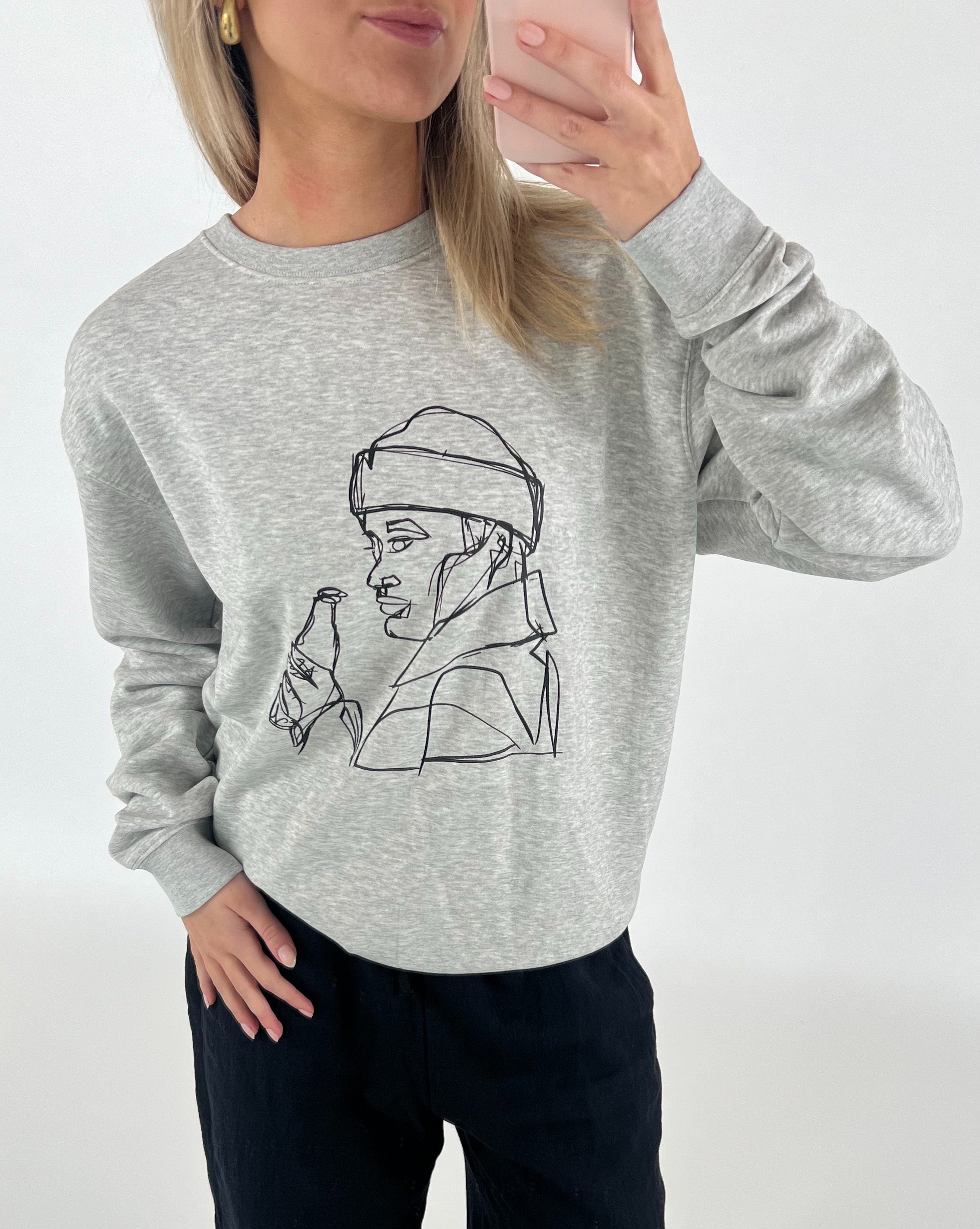 Person iført grå sweatshirt med lineart-tegning af en person med flaske. Personen holder en mobiltelefon foran et neutralt bagtæppe.