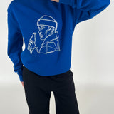 Blå sweatshirt med line art af en person med en flaske. Personen holder en hånd nær ansigtet. Baggrunden er neutral.