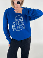En person iført en blå sweatshirt med en hvid, stiliseret tegning af en person der holder en flaske, tager et spejlbillede i et lyst rum.