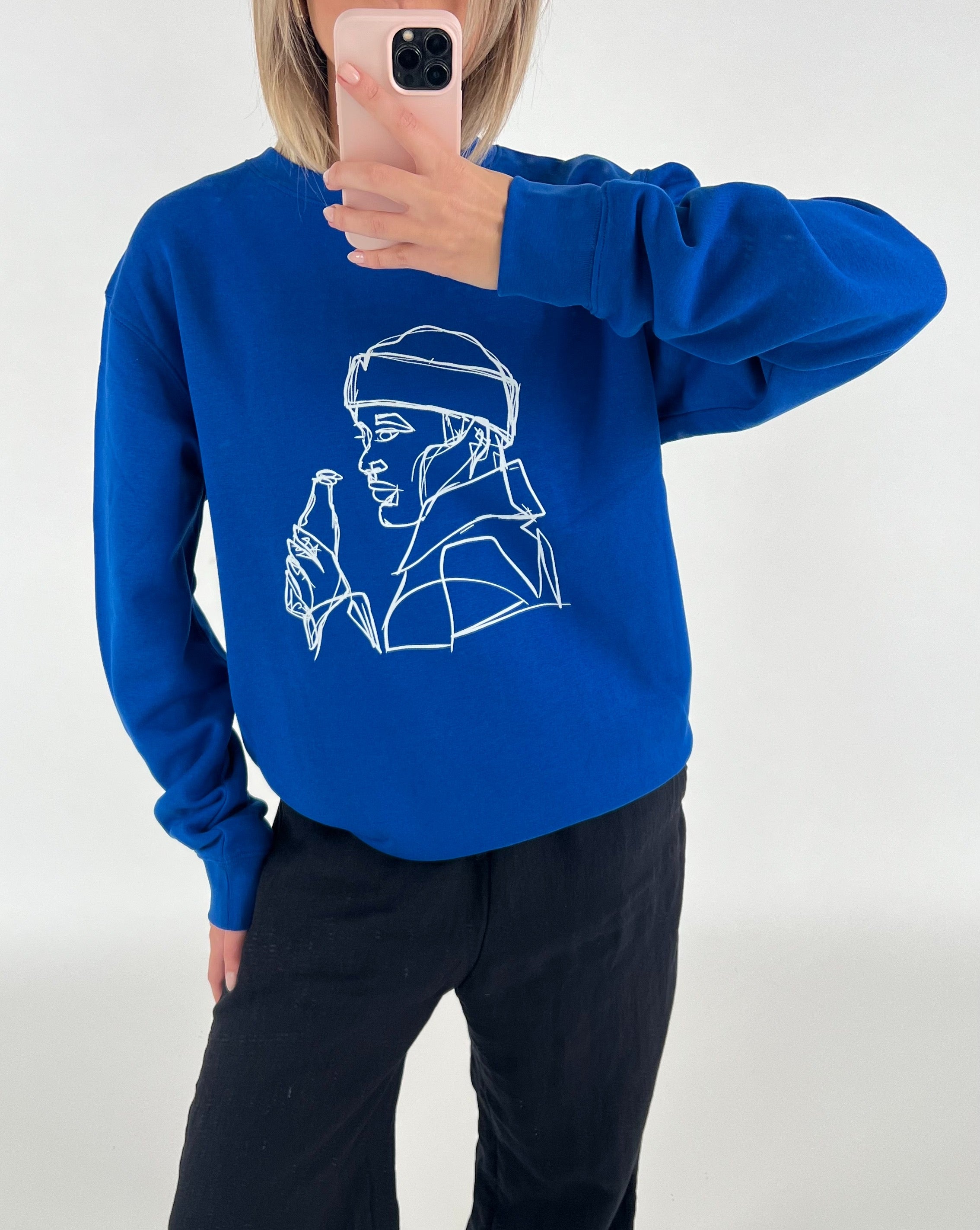 En person iført en blå sweatshirt med en hvid, stiliseret tegning af en person der holder en flaske, tager et spejlbillede i et lyst rum.