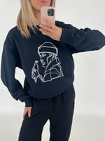 En person tager et spejlbillede iført en sort sweatshirt med en line-tegning af en person med en drikkeflaske. Baggrunden er neutral og lys.