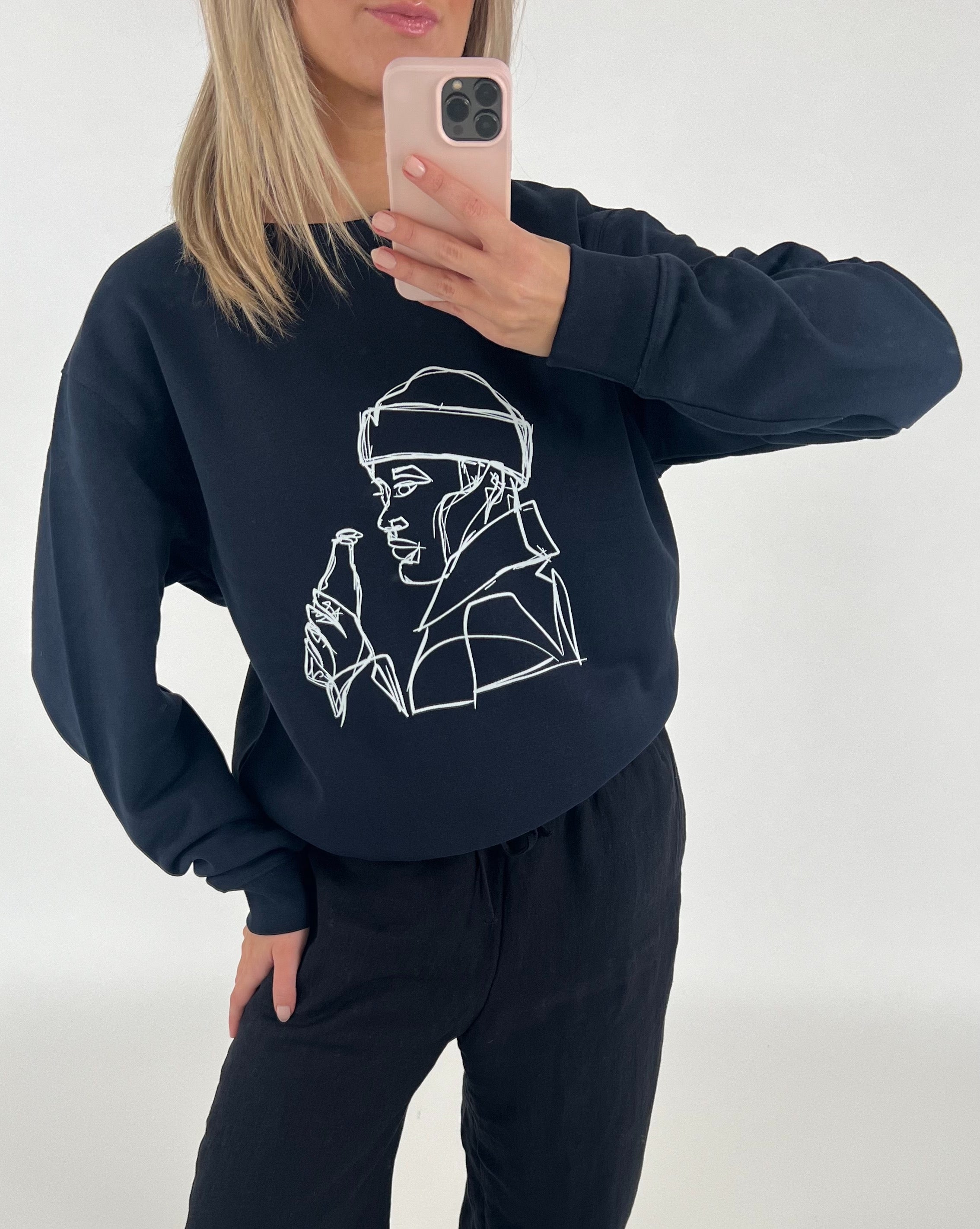 En person tager et spejlbillede iført en sort sweatshirt med en line-tegning af en person med en drikkeflaske. Baggrunden er neutral og lys.