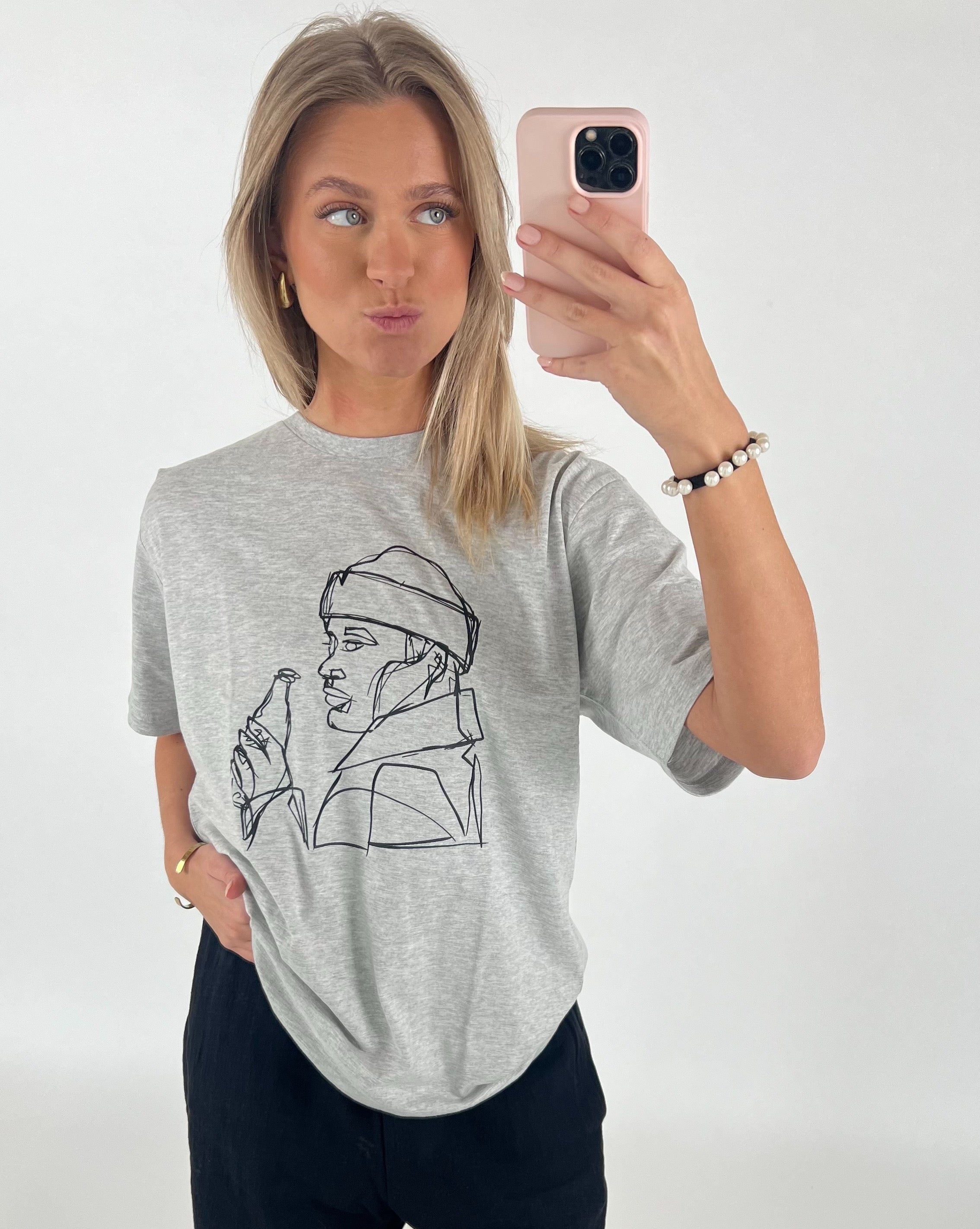 En kvinde fotograferer sig selv med en smartphone, iført en grå T-shirt med en lineart-tegning af en person, der holder en flaske. Hun står i et lyst rum.