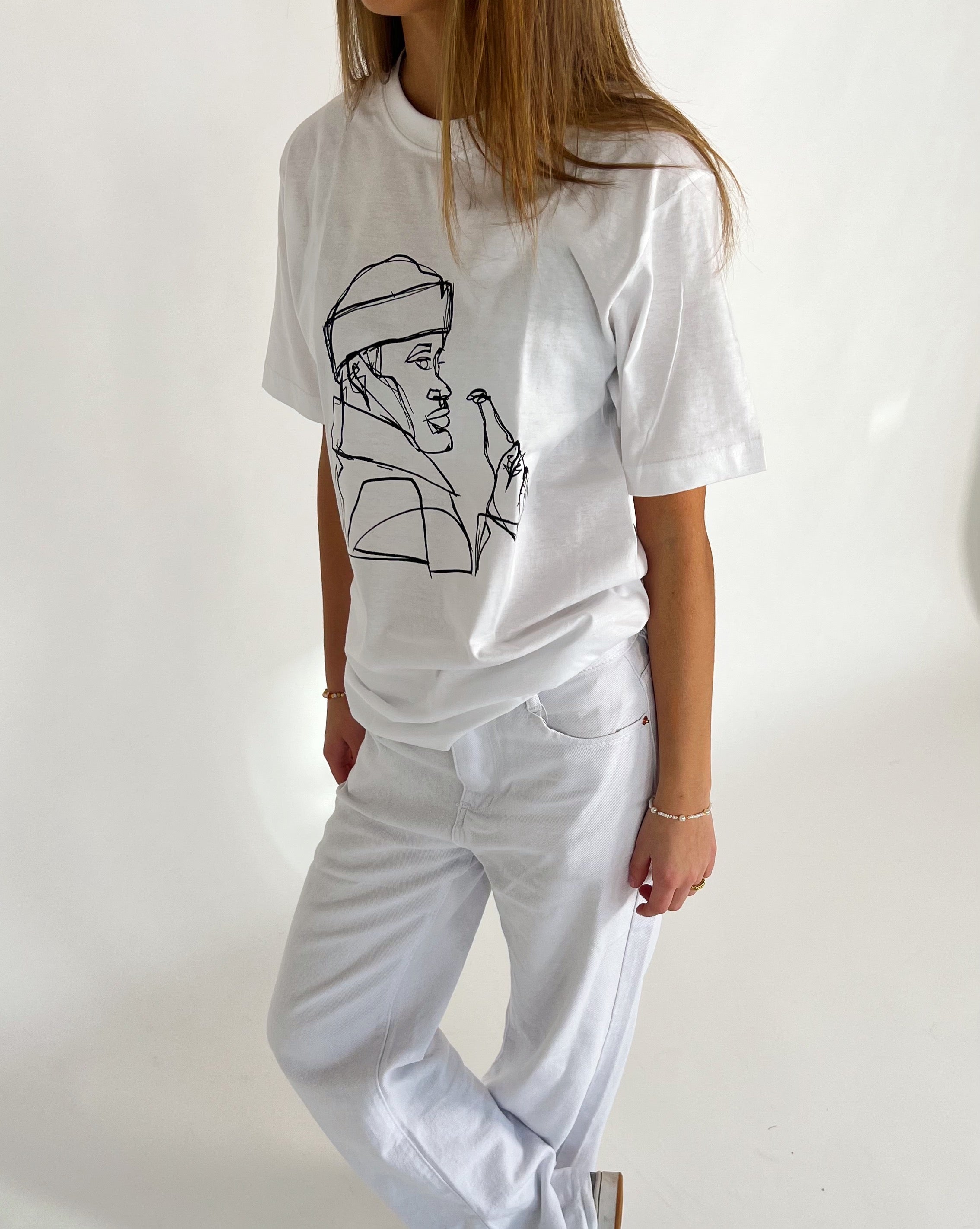 En person iført hvid t-shirt med en sort linjetegning af en person på forsiden. Personen står afslappet, iført hvide bukser, mod en lys baggrund.