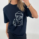 Kvinde i sort t-shirt med line art-tryk af person, holder mobiltelefon op til spejl. Hun står i minimalistisk, hvidt rum iført perlearmbånd.