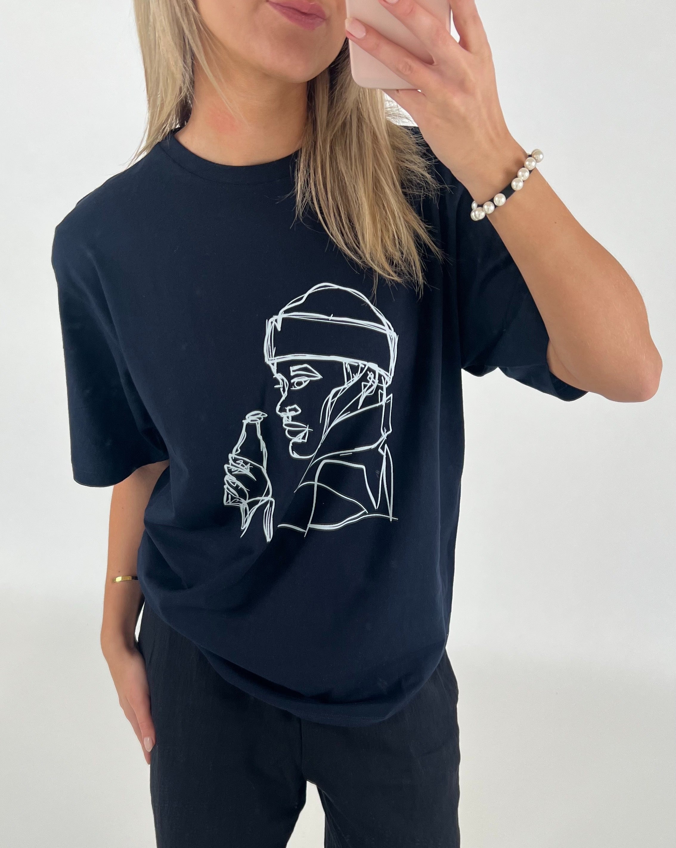 Kvinde i sort t-shirt med line art-tryk af person, holder mobiltelefon op til spejl. Hun står i minimalistisk, hvidt rum iført perlearmbånd.