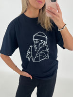 Kvinde iført sort T-shirt med en hvidt lineart design af en person med hætte og flaske. Hun ser sig i spejlet med en mobiltelefon i et lyst rum.