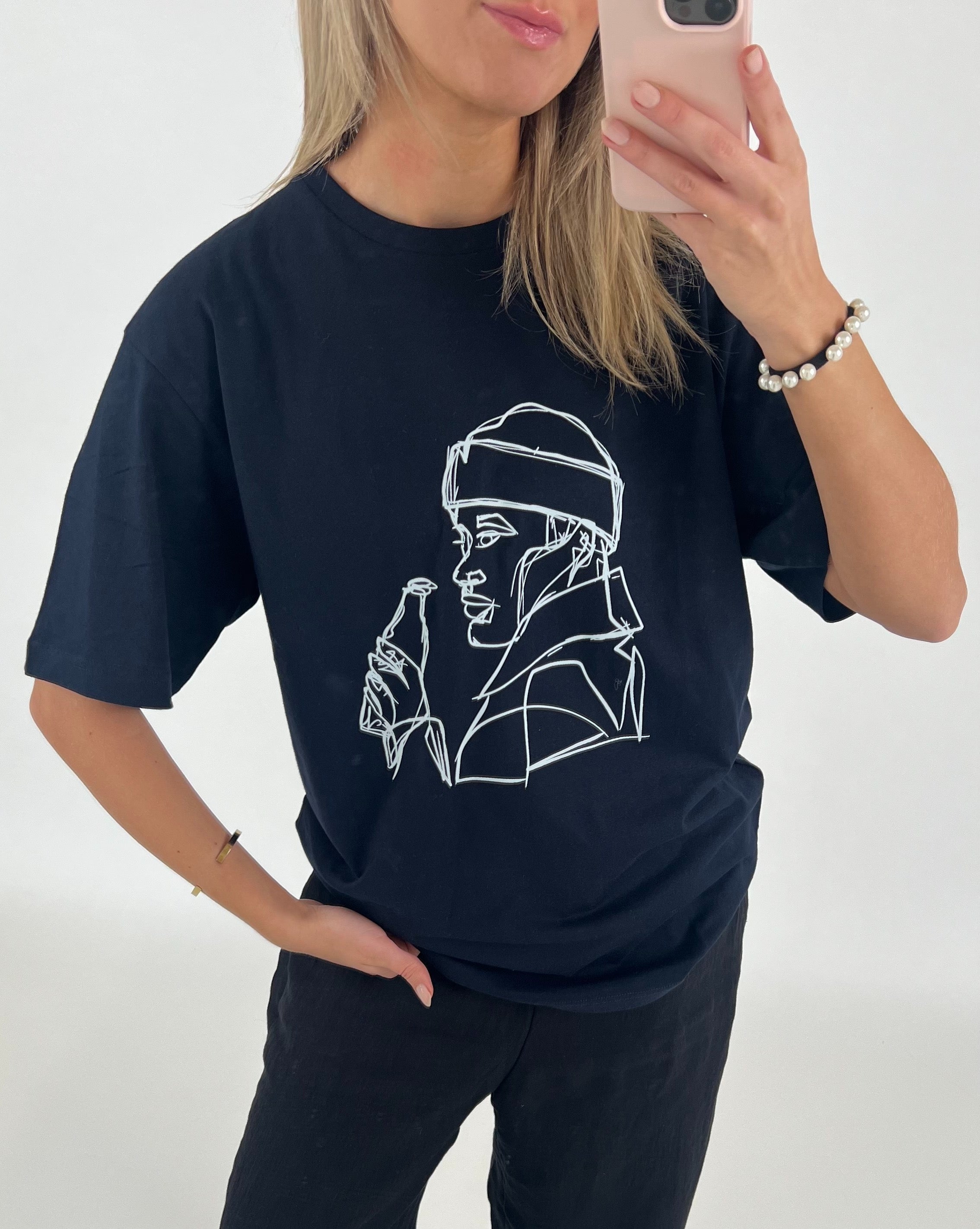 Kvinde iført sort T-shirt med en hvidt lineart design af en person med hætte og flaske. Hun ser sig i spejlet med en mobiltelefon i et lyst rum.