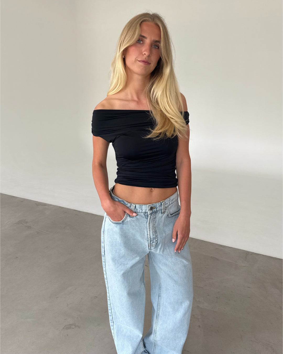 Modelfoto af pige som bærer sort THORA offshoulder top, og lyseblå jeans . Alt er fra BySofieSønderby