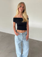 Modelfoto af pige som bærer sort THORA offshoulder top, og lyseblå jeans . Alt er fra BySofieSønderby