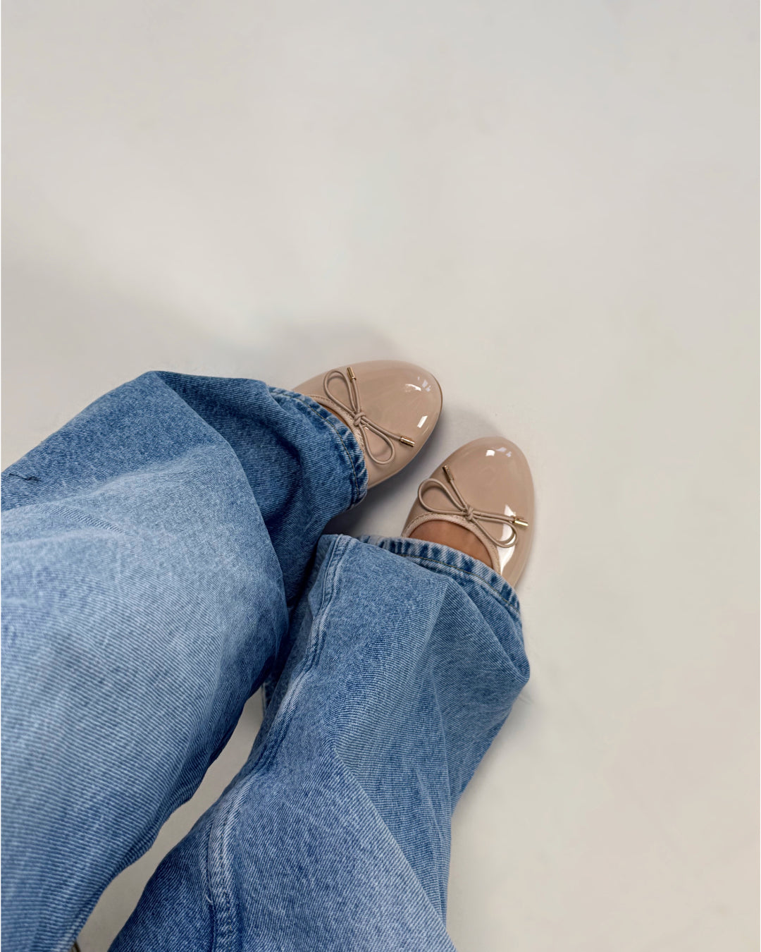 Brune ballerinasko med sløjfer bæres af en person iført lyseblå jeans. Personen står på et lyst gulv.