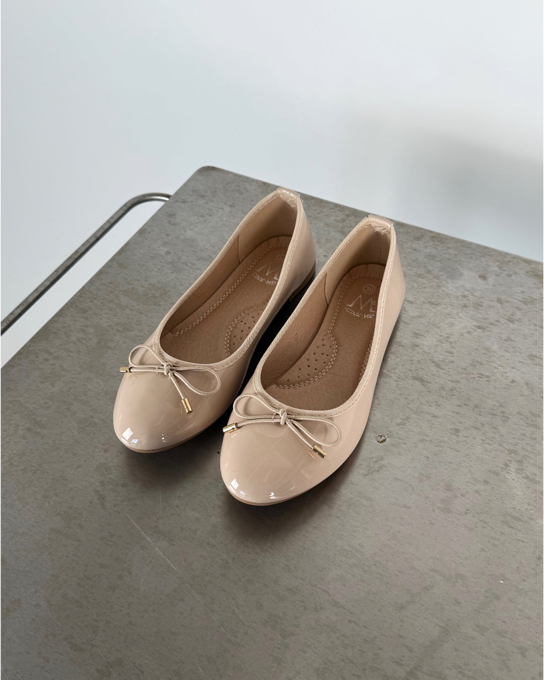Beige ballerinasko med sløjfe ligger stille på en gråmetal overflade. Indersålen bærer teksten "M" og "38". En minimalistisk, lys baggrund omkranser dem.