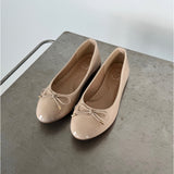 Beige ballerinasko med sløjfe ligger stille på en gråmetal overflade. Indersålen bærer teksten "M" og "38". En minimalistisk, lys baggrund omkranser dem.