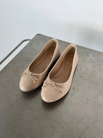 Beige ballerinasko med sløjfe ligger stille på en gråmetal overflade. Indersålen bærer teksten "M" og "38". En minimalistisk, lys baggrund omkranser dem.