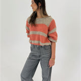 En kvinde står og kigger til siden. Hun bærer en stribet trøje i orange og beige samt grå jeans. Baggrunden er hvid og minimalistisk.