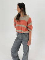 En kvinde står og kigger til siden. Hun bærer en stribet trøje i orange og beige samt grå jeans. Baggrunden er hvid og minimalistisk.