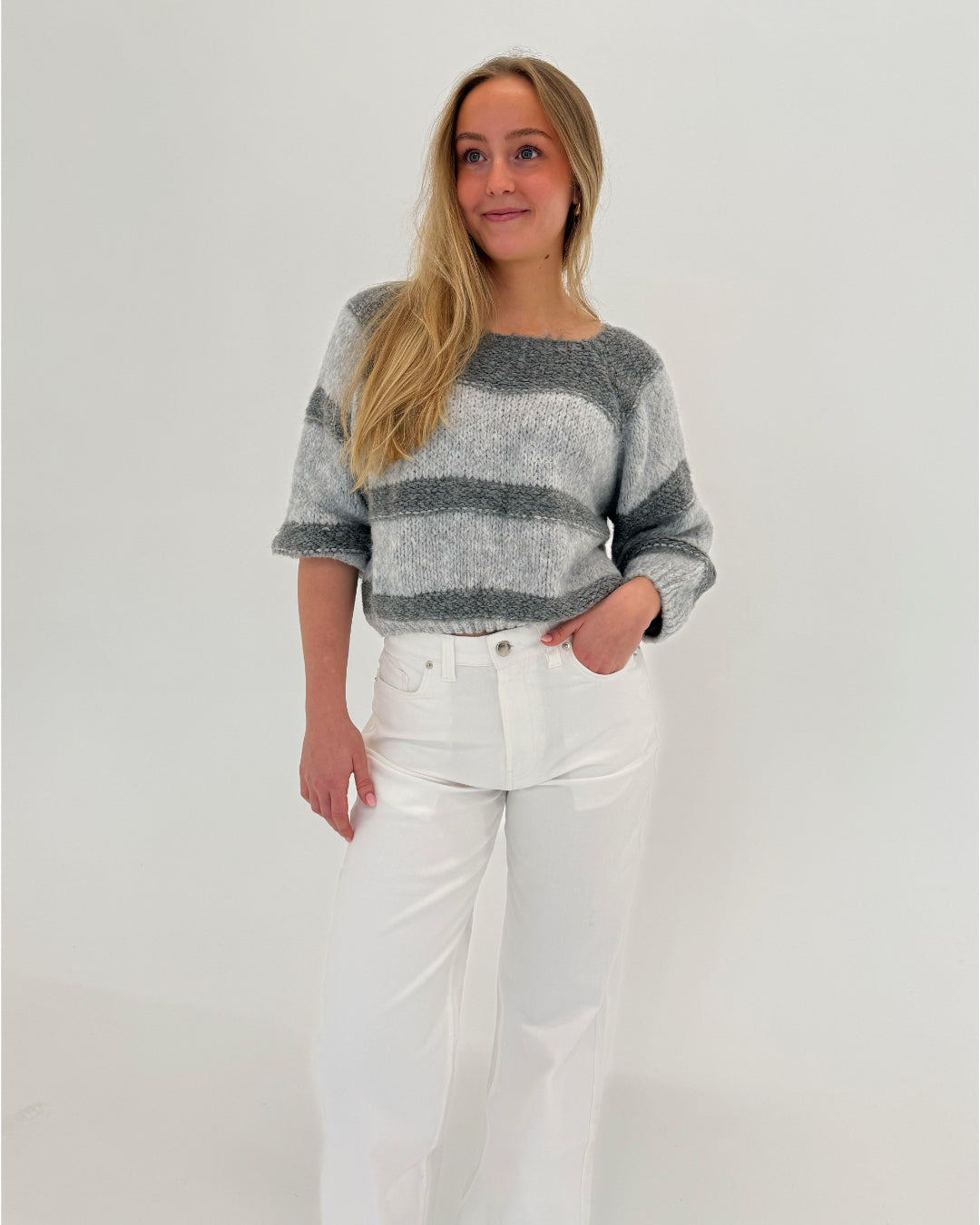 En person iført en gråstribet sweater og hvide bukser står afslappet med en hånd i lommen mod en lys baggrund.