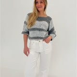 En person iført en gråstribet sweater og hvide bukser står afslappet med en hånd i lommen mod en lys baggrund.