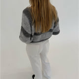 En person med langt hår står med ryggen til, iført en gråstribet sweater og hvide bukser, i et minimalistisk, lyst rum.