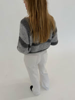 En person med langt hår står med ryggen til, iført en gråstribet sweater og hvide bukser, i et minimalistisk, lyst rum.