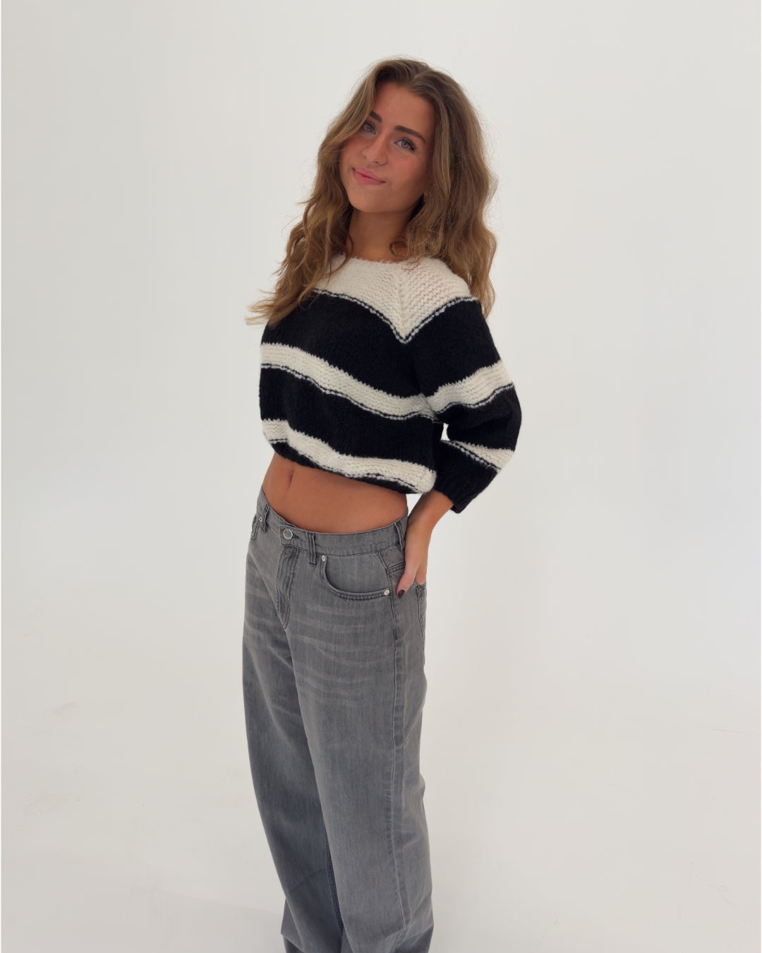 Kvinde i en sort-hvid stribet sweater og grå jeans står afslappet med hånden i lommen, smilende i et lyst rum med en hvid baggrund.