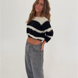 Kvinde i en sort-hvid stribet sweater og grå jeans står afslappet med hånden i lommen, smilende i et lyst rum med en hvid baggrund.