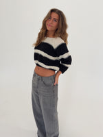 Kvinde i en sort-hvid stribet sweater og grå jeans står afslappet med hånden i lommen, smilende i et lyst rum med en hvid baggrund.