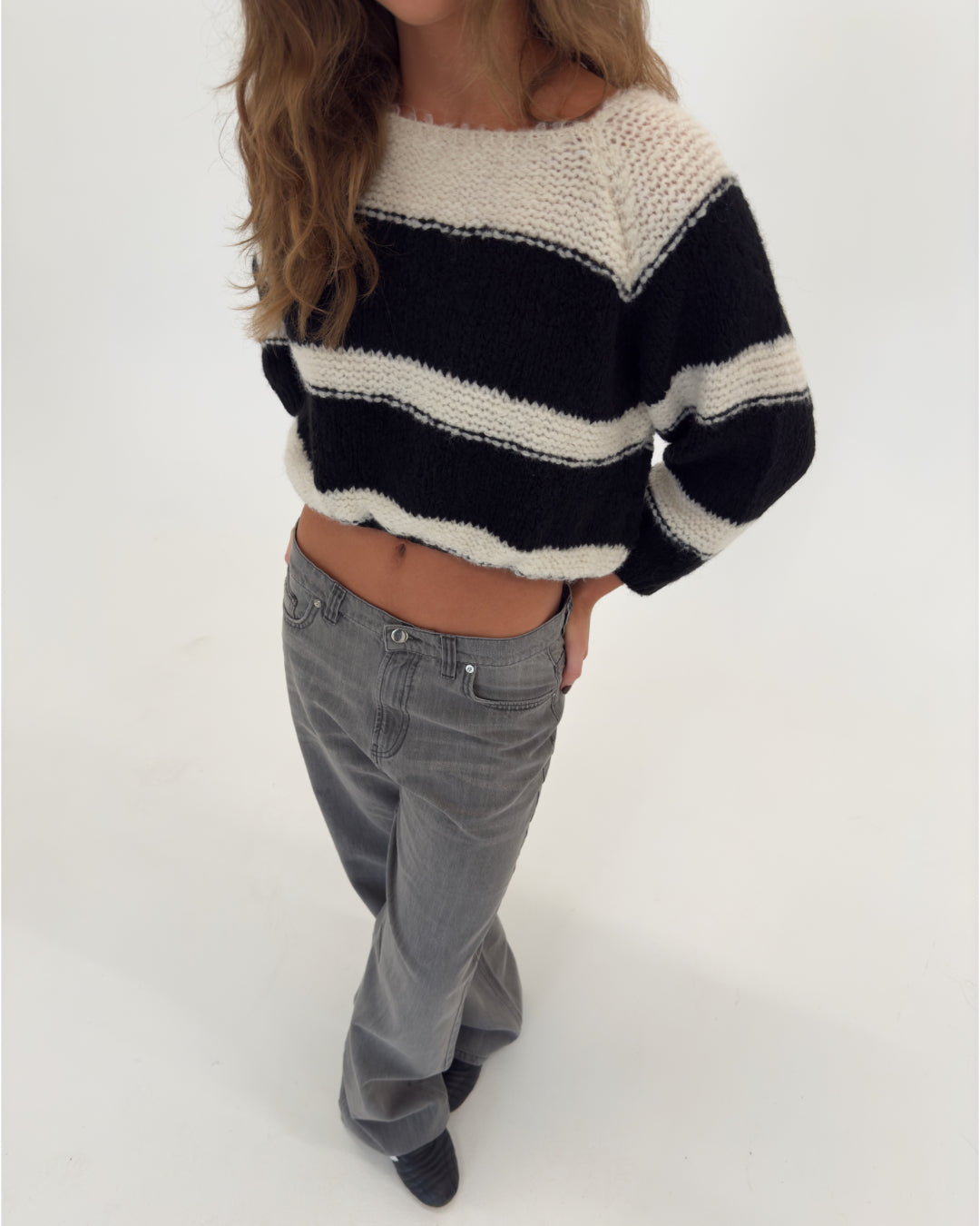 En person iført en sort-hvid stribet sweater står med hænderne i lommerne på et par grå bukser i et lyst studie.