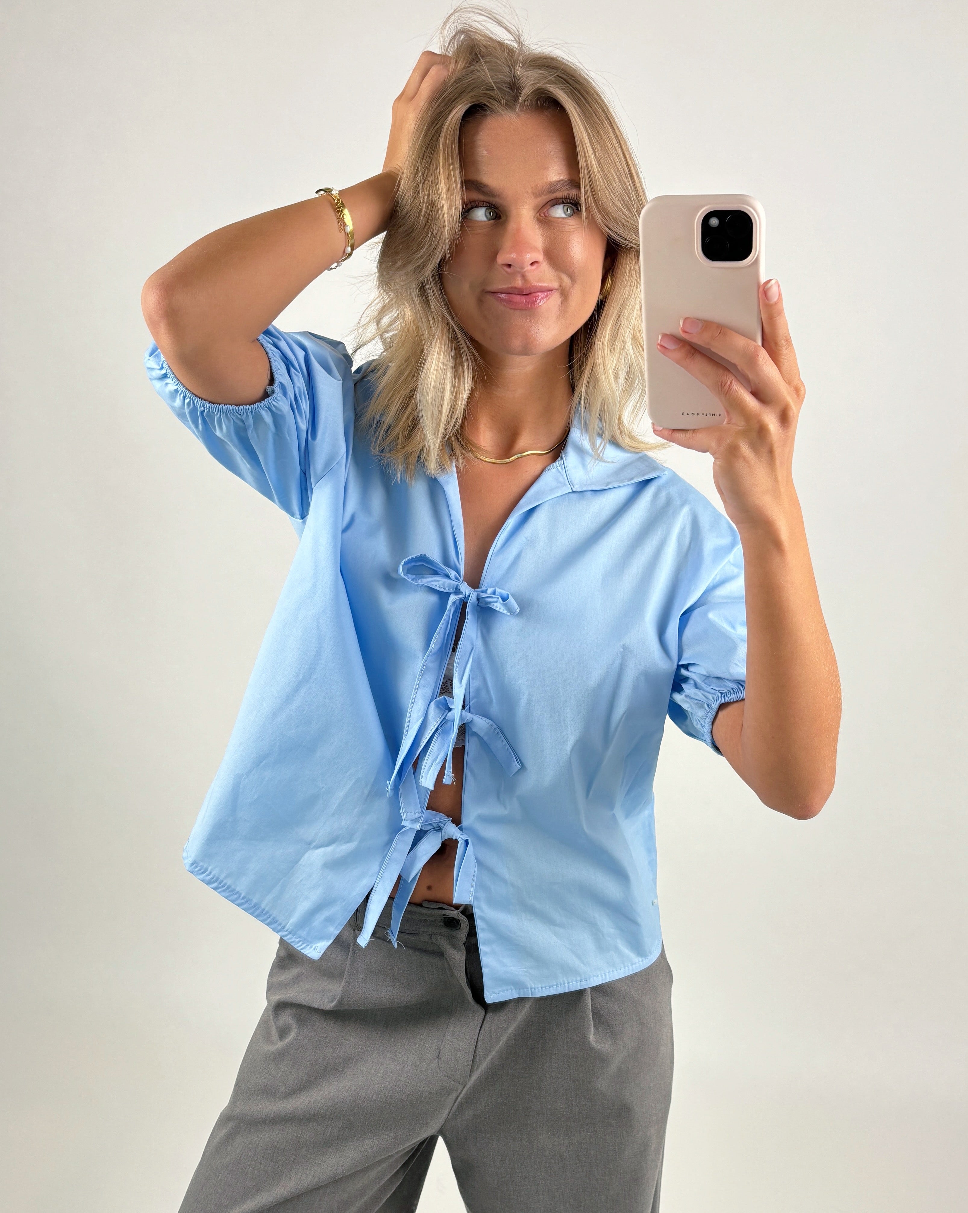 En person iført en blå bluse med sløjfer tager en selfie med en smartphone. Personen står med hånden i håret i en neutral indstilling.