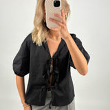 En person iført sort skjorte tager et selfie med en mobiltelefon, ansigtet dækket af telefonen. Personen står i et neutralt, lyst rum.
