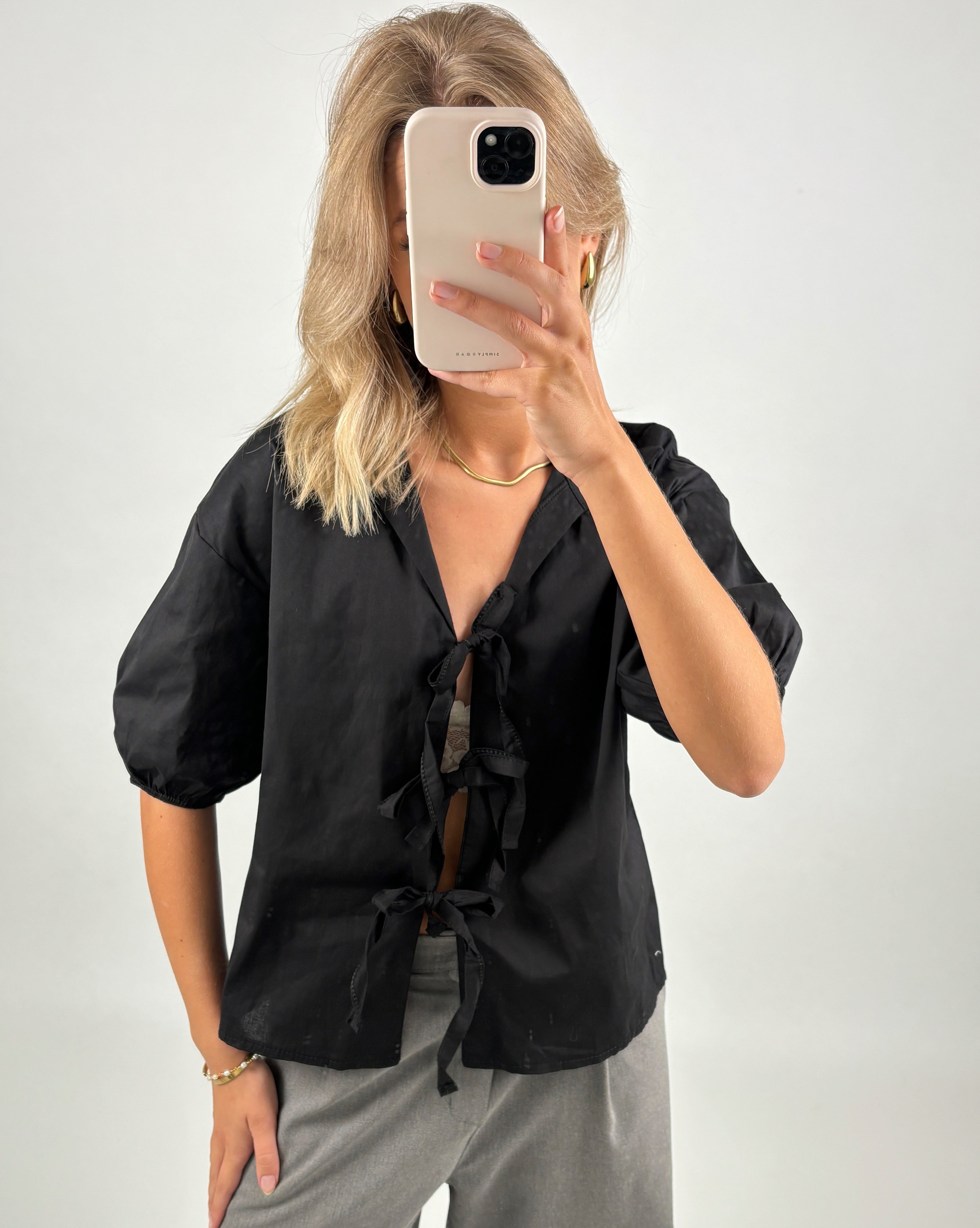 En person iført sort skjorte tager et selfie med en mobiltelefon, ansigtet dækket af telefonen. Personen står i et neutralt, lyst rum.
