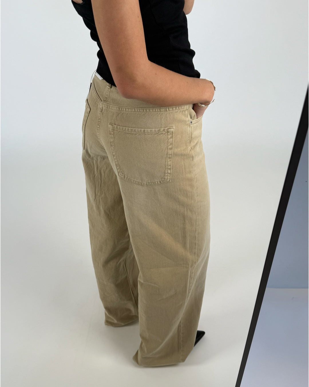 En person iført beige bukser står med hænderne i lommerne. Personen har en sort tanktop på, og baggrunden er hvid og minimalistisk.