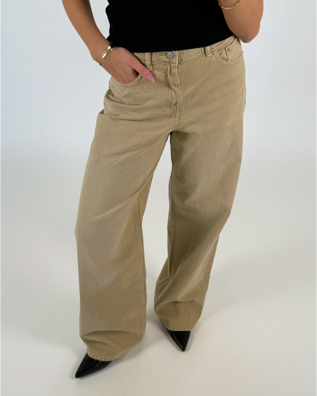 Beige jeans bæres af en person iført en sort top, der står med hænderne i lommerne. Baggrunden er lys, og sorte sko er synlige.