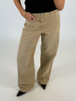 Beige jeans bæres af en person iført en sort top, der står med hænderne i lommerne. Baggrunden er lys, og sorte sko er synlige.