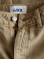 Knappen på et par beige bukser med lynlås er åben. Et mærke med teksten "JJXX" er synligt på indersiden.