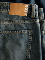 Et par blå jeans ligger fladt, med en brun læderlignende lap påsyet i taljen. På lappen står teksten "LXX". Jeansene har en tydelig synlig baglomme.