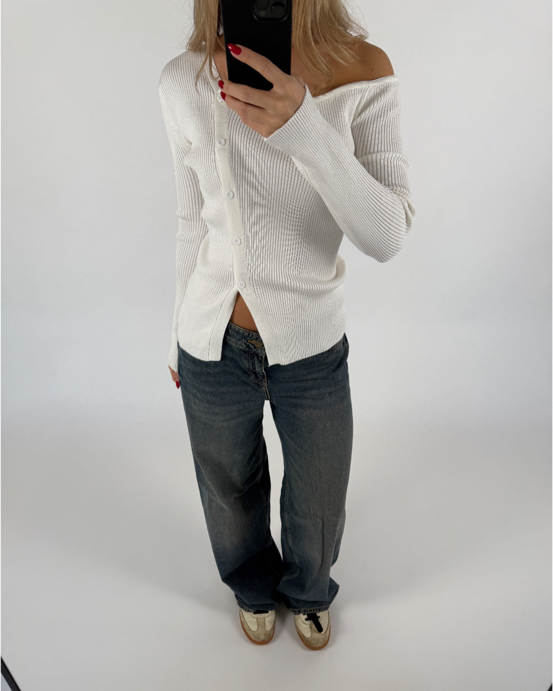 En person iført en hvid, off-shoulder cardigan tager et spejlbillede med en mobiltelefon. De har mørke jeans og sko på en lys baggrund.