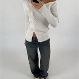 En person iført en hvid, off-shoulder cardigan tager et spejlbillede med en mobiltelefon. De har mørke jeans og sko på en lys baggrund.