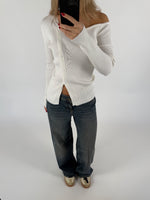En person iført en hvid, off-shoulder cardigan tager et spejlbillede med en mobiltelefon. De har mørke jeans og sko på en lys baggrund.