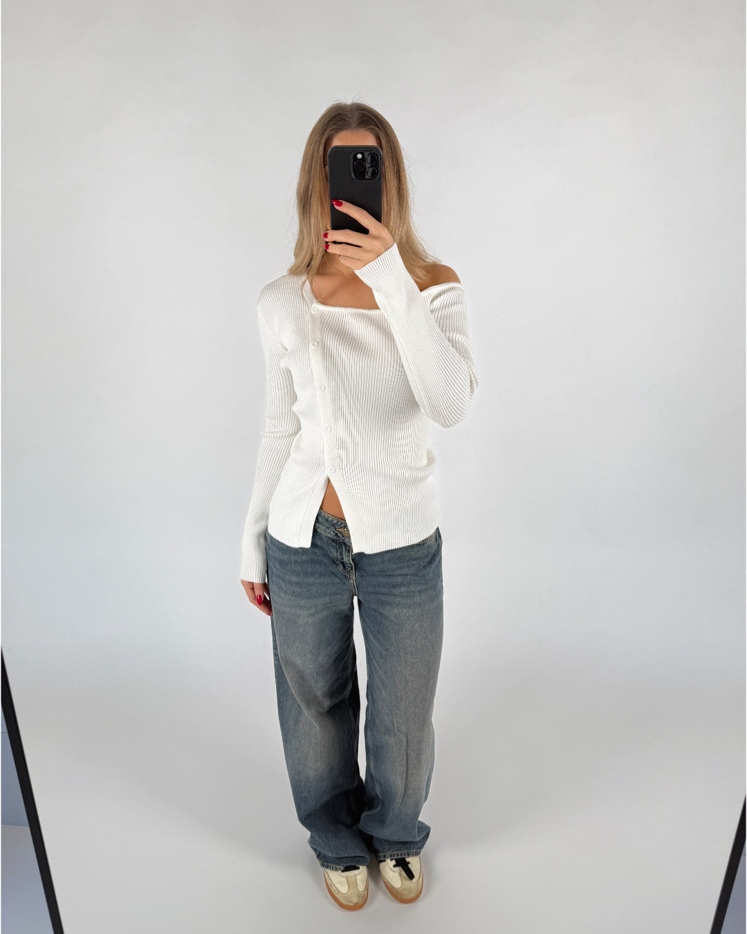 En person tager et spejlbillede iført en hvid sweater og løse jeans, stående i et rum med en lys baggrund. Personen holder en sort telefon, som skjuler ansigtet.