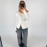 En person tager et spejlbillede iført en hvid sweater og løse jeans, stående i et rum med en lys baggrund. Personen holder en sort telefon, som skjuler ansigtet.
