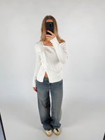 En person tager et spejlbillede iført en hvid sweater og løse jeans, stående i et rum med en lys baggrund. Personen holder en sort telefon, som skjuler ansigtet.
