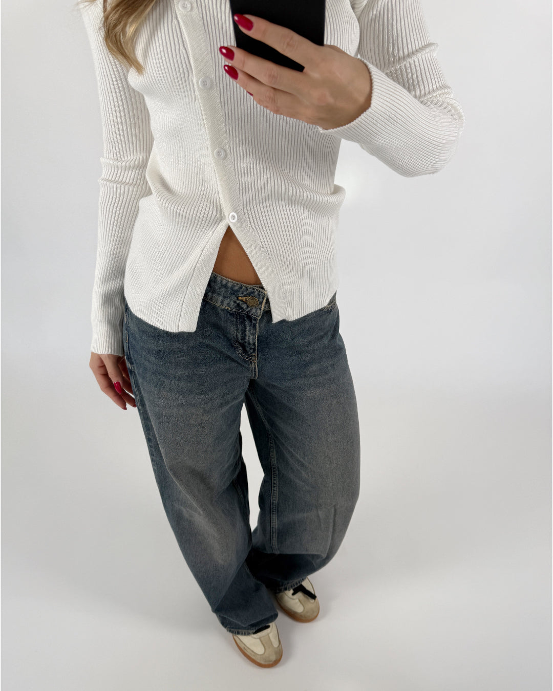 En person bærer en hvid striktrøje og løstsiddende jeans, fotograferer sig selv med en smartphone i et neutralt rum. Personen har rød neglelak på.