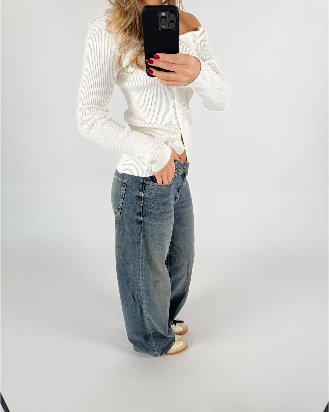 En person tager en selfie iført en hvid striktrøje og løse jeans. Telefonen dækker ansigtet, og omgivelserne er en simpel lysegrå baggrund.