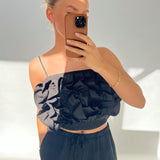 Person tager selfie, iført sort top med flæser, holder brun telefon foran ansigtet i lyst rum. Arm bærer gyldent armbånd. Ingen tekst til stede.