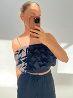 Person tager selfie, iført sort top med flæser, holder brun telefon foran ansigtet i lyst rum. Arm bærer gyldent armbånd. Ingen tekst til stede.